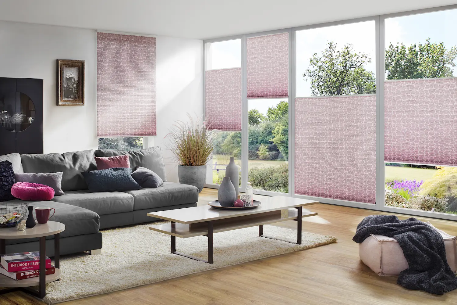 Modernes Wohnzimmer mit stilvollen rosa Plissees, grauem Sofa, dekorativen Kissen und grossen Fenstern mit Blick ins Grüne.