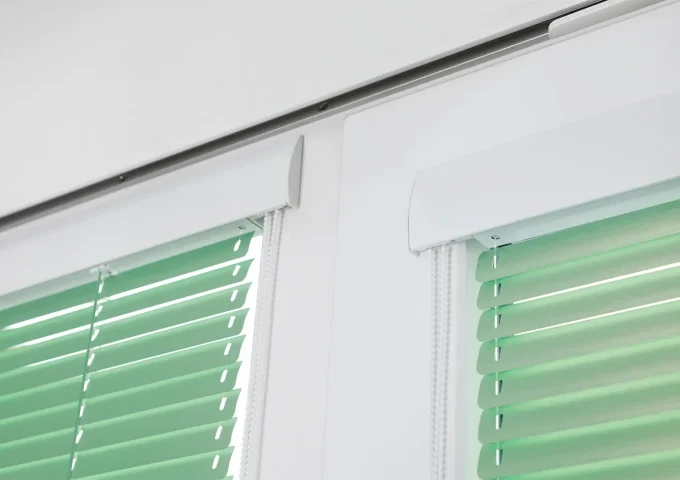 Zwei nebeneinanderliegende Fenster mit hellgrünen Lamellen und weissen Abdeckungen an der Oberseite.