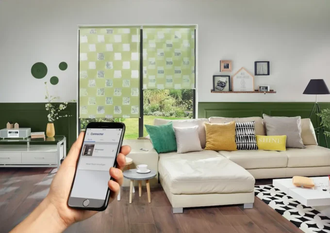 Ein heller Wohnraum mit gemütlicher Sitzecke, Blick in den Garten und einer Hand mit Smartphone im Vordergrund.