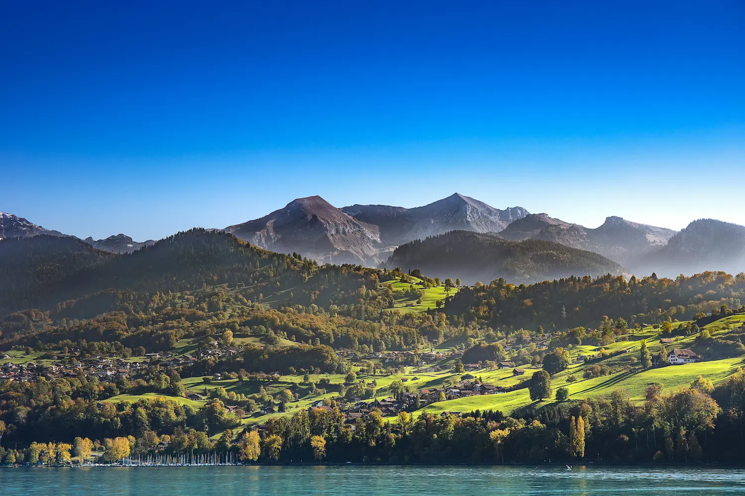 Grüne Hügellandschaft mit Dörfern vor einer imposanten Bergkulisse und einem klaren See